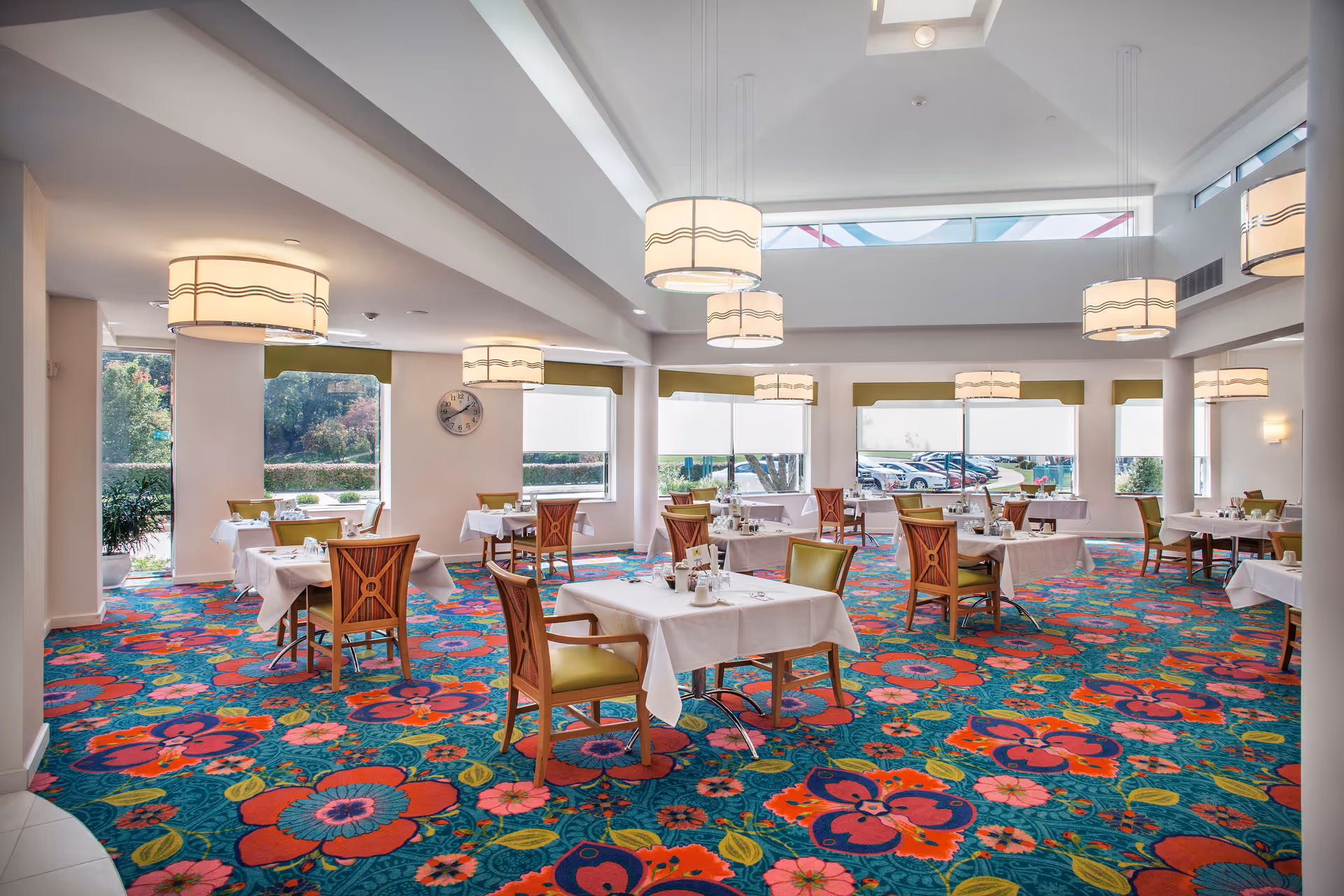 Bright dining room with multiple tables covered in white tablecloths, each set with chairs. The room features large windows letting in natural light, colorful floral carpet, and modern hanging light fixtures.