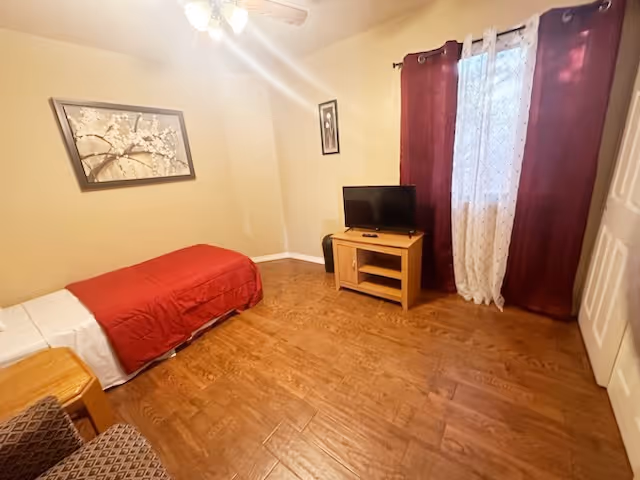 A small bedroom with a single bed covered with a red blanket and white sheets, a wooden nightstand, a small TV on a wooden stand, a window with sheer white and dark red curtains, a ceiling fan with lights, and two framed pictures on the beige walls. The floor is wooden.