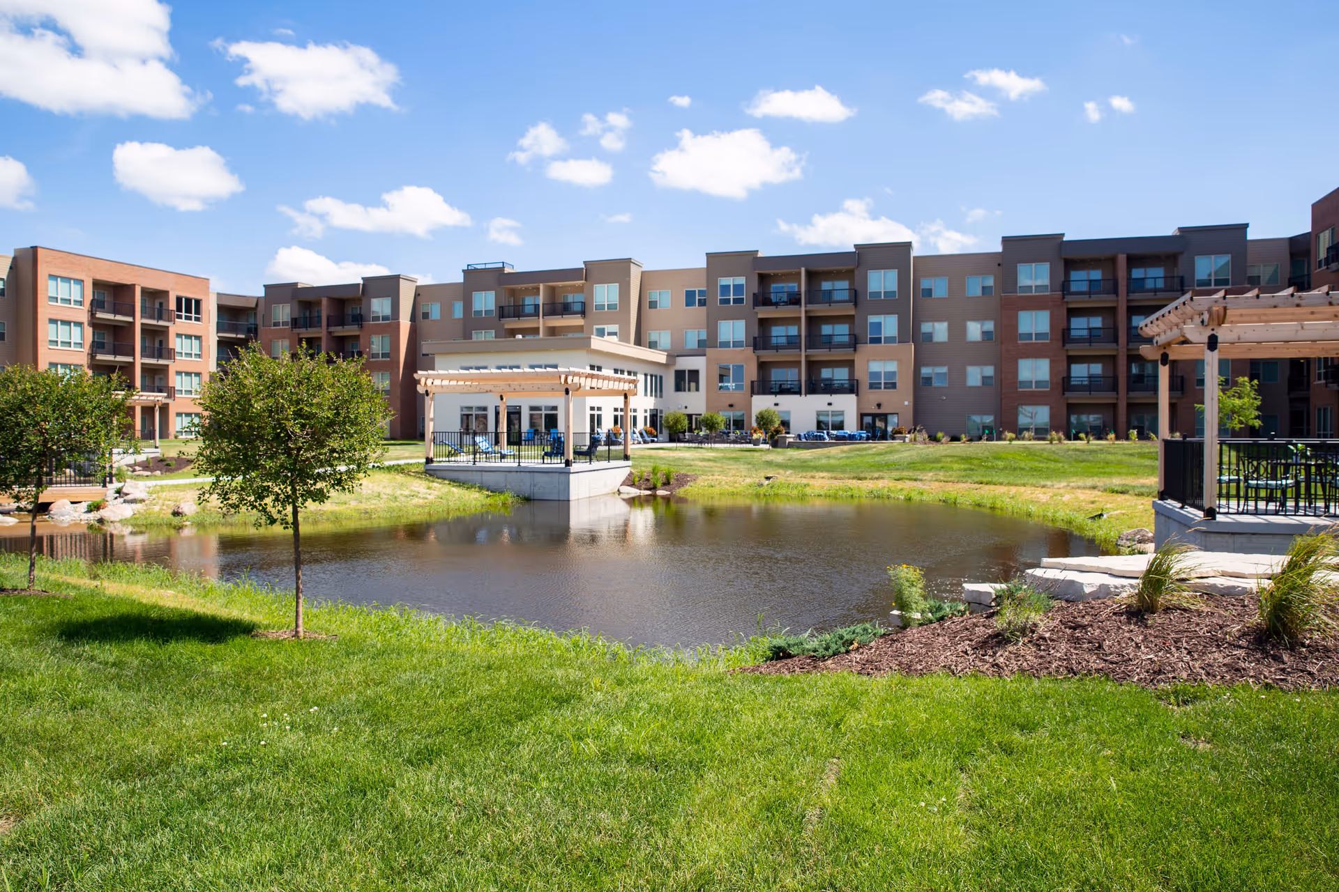 View of a senior living facility named Attivo Trail Waukee with a pond in the foreground, green grass, small trees, and a large multi-story building with balconies in the background under a blue sky with scattered clouds.