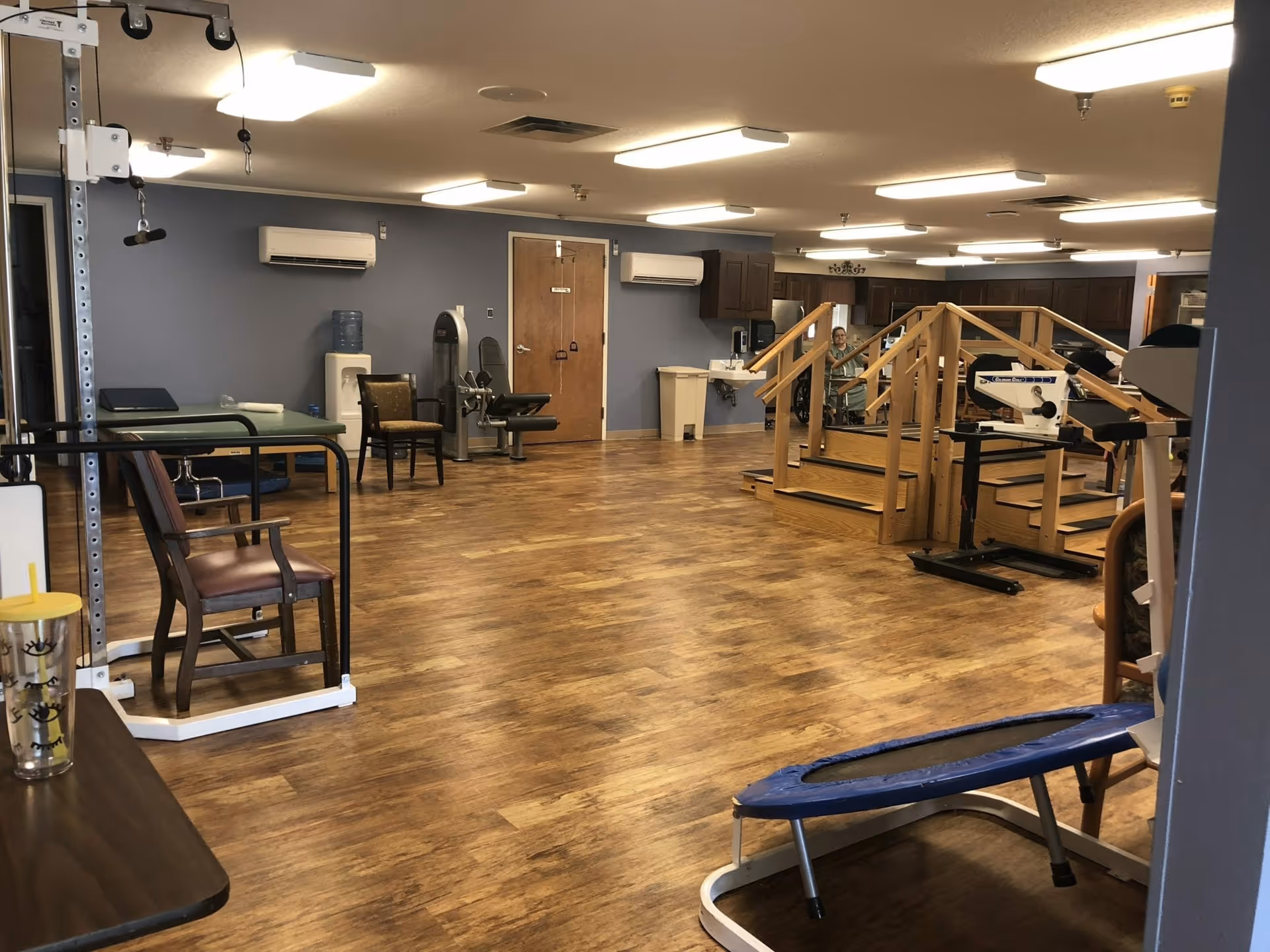 Rehabilitation gym room with wooden floors, exercise equipment, therapy stairs and chairs.