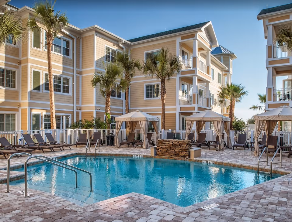 Swimming pool courtyard with lounge chairs, cabanas, palm trees, and multi-story yellow residential buildings.