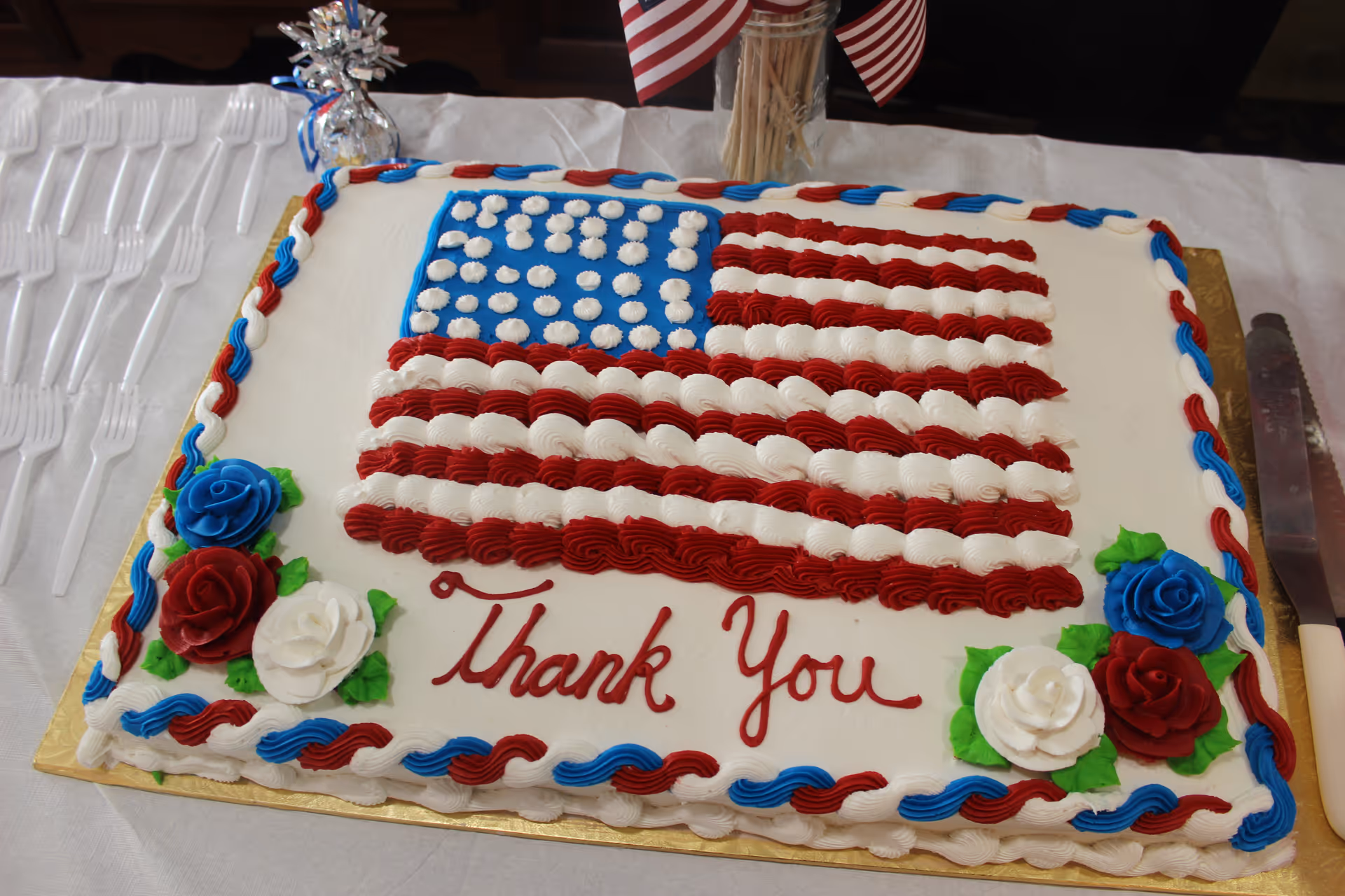 Sheet cake decorated as an American flag with "Thank You" icing and red, white, and blue rose decorations on a table.