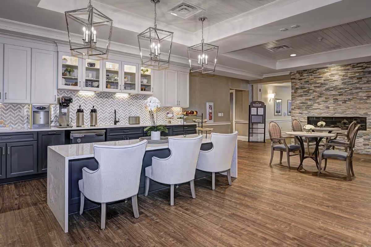 Open common area with a kitchen island, upholstered chairs, pendant lights and dining tables in a senior living facility.