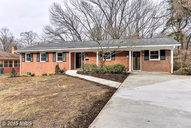 Single-story brick residential building with a gray shingled roof, multiple windows with black shutters, two front doors, a concrete driveway, and a curved concrete walkway leading to the entrance. The yard has sparse grass and some small shrubs near the building. Leafless trees are visible in the background.