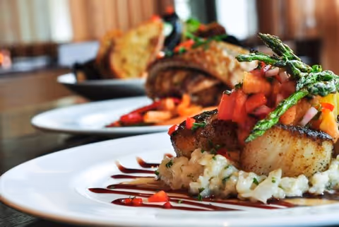 Close-up of a gourmet dish featuring seared scallops on a bed of risotto, topped with diced tomatoes and asparagus, with another plated dish and bread in the background on a dining table.