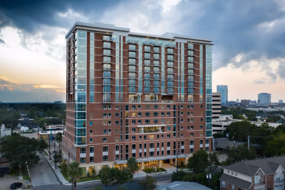 A tall multi-story brick and glass building at dusk with lights on inside, surrounded by trees and smaller buildings in an urban area under a cloudy sky.