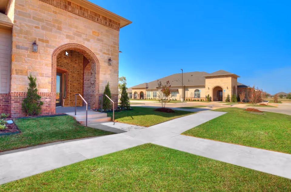 Exterior view of a senior living facility with stone and brick buildings, well-maintained green lawns, and concrete walkways under a clear blue sky.