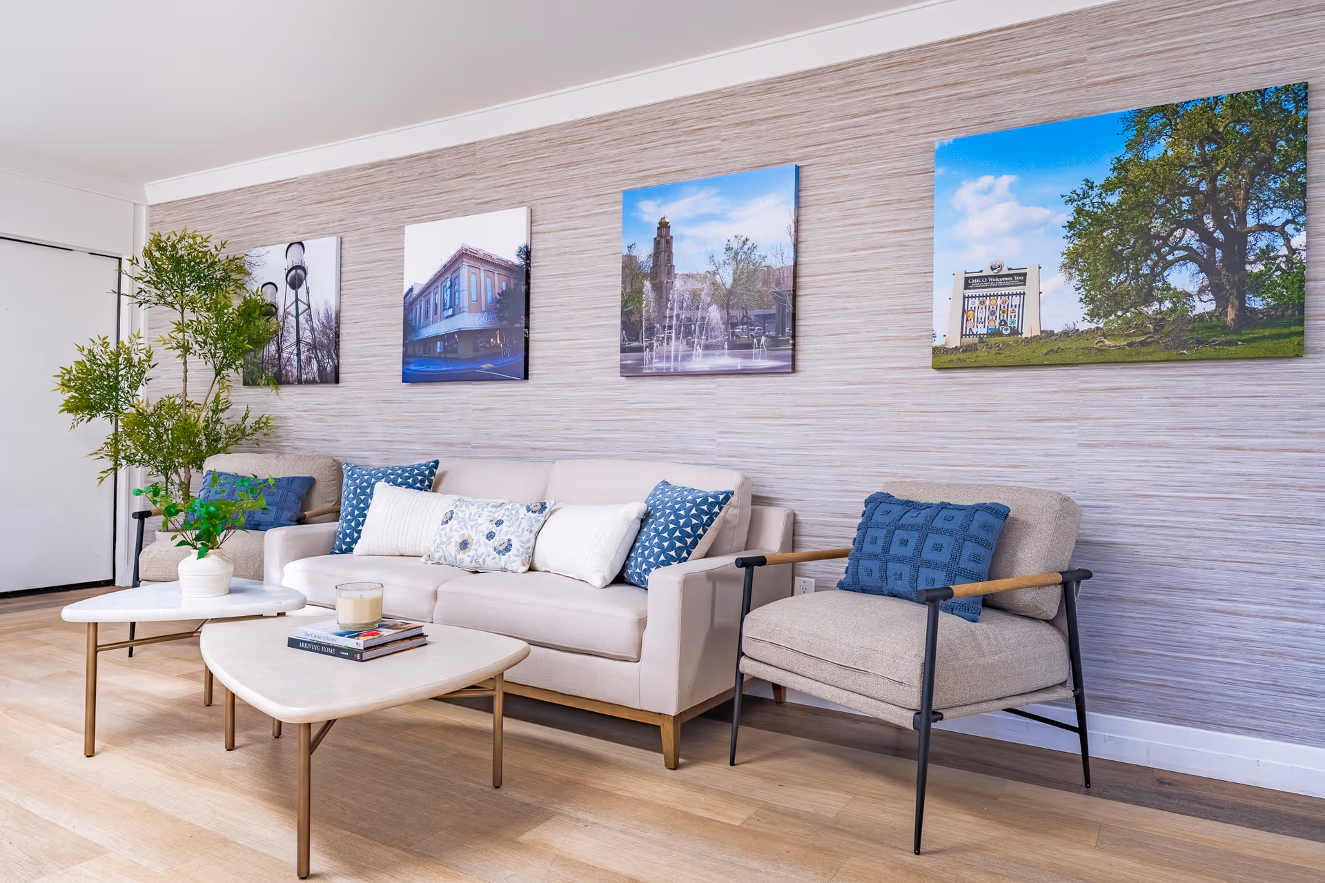 A cozy living room area with a beige sofa adorned with blue and white patterned cushions, a matching armchair with a blue cushion, two modern white coffee tables with gold legs, a potted plant on one table, and framed photographs of outdoor scenes hanging on a textured beige wall.