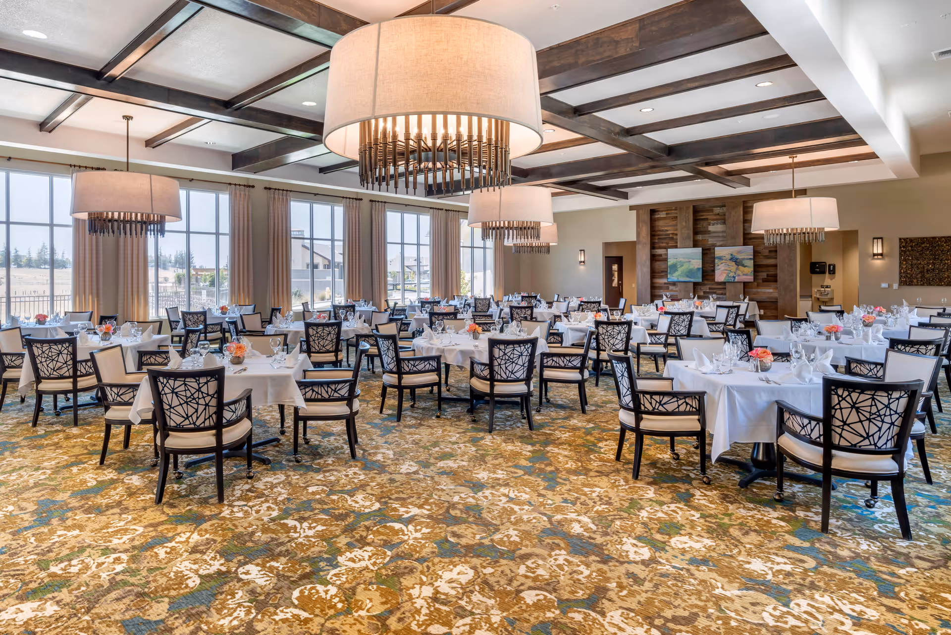 A spacious dining room with multiple tables covered in white tablecloths, each set with glassware, napkins, and small floral centerpieces. The room features large windows with beige curtains allowing natural light to fill the space, decorative ceiling beams, and large modern chandeliers hanging from the ceiling. The carpet has a patterned design in shades of brown, blue, and beige.