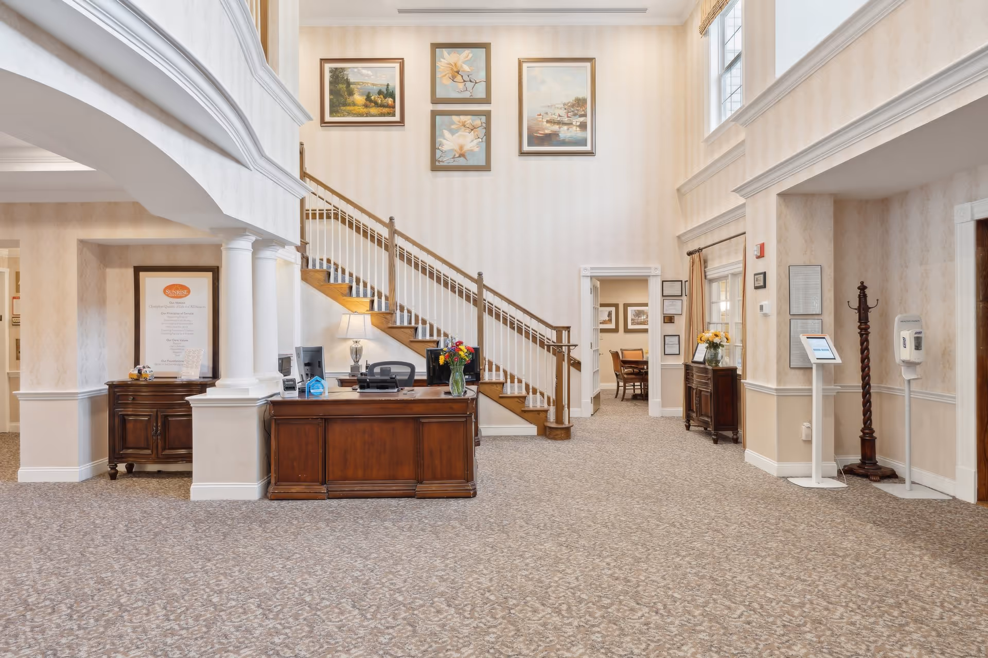 Interior view of a senior living facility lobby with a wooden reception desk, a staircase with wooden handrails, framed paintings on the wall, and a carpeted floor. There are columns and archways, a coat rack, hand sanitizer dispenser, and a small table with flowers and a digital kiosk.
