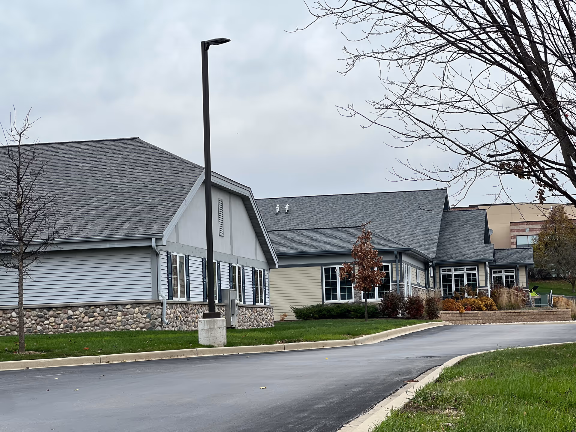 Front exterior of a single-story senior living building with a stone base, driveway, lamp post, and bare trees under a cloudy sky.