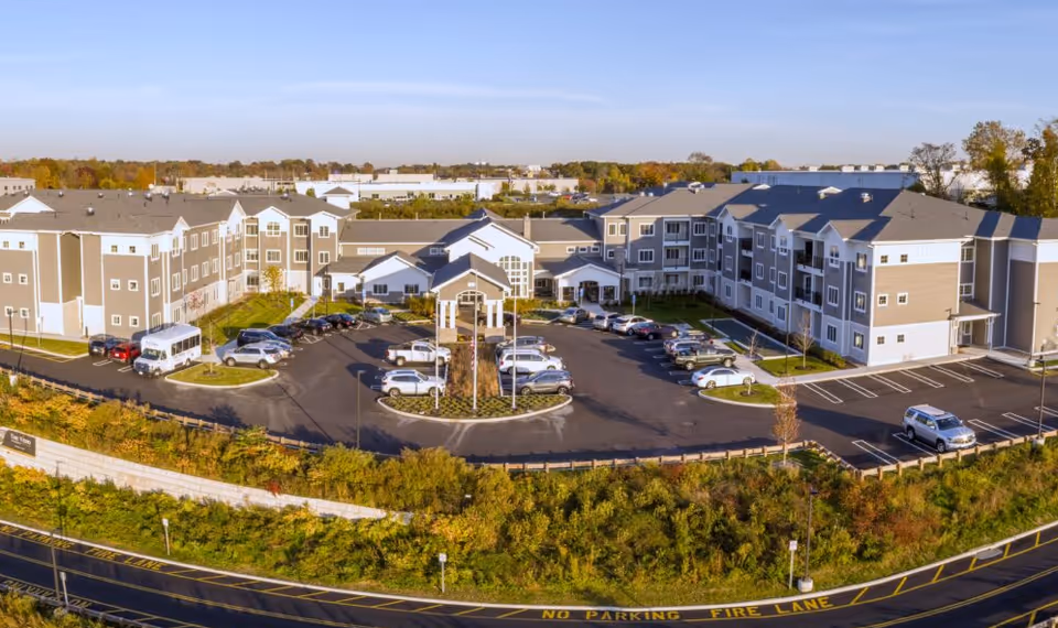 Aerial view of a large senior living facility with multiple connected buildings surrounding a central parking lot. The buildings are three stories high with beige and white exterior walls and gray roofs. Several cars are parked in the lot, and there is greenery and trees around the property. The sky is clear and blue.
