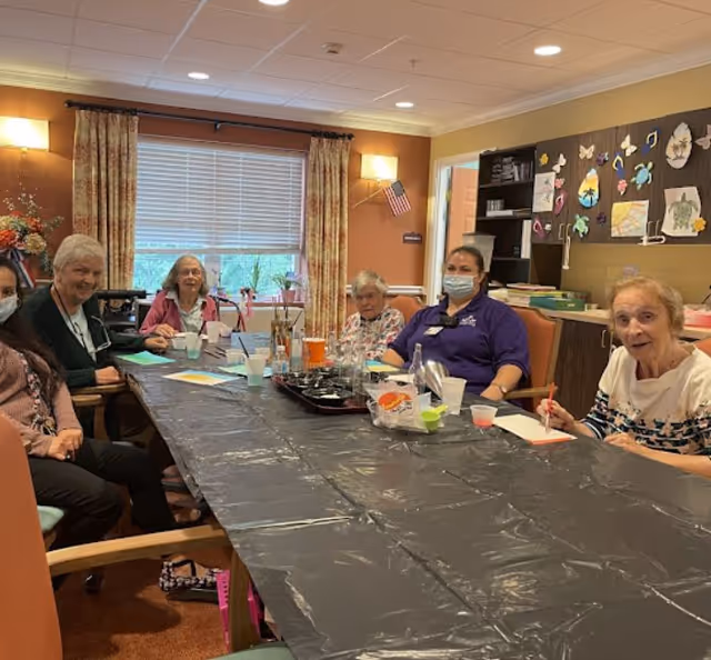 A group of elderly women and a caregiver wearing a mask sitting around a long table covered with a black plastic tablecloth in a well-lit room. The table has art supplies and cups on it. The room has warm-colored walls, a window with curtains, and a bulletin board with colorful artwork.