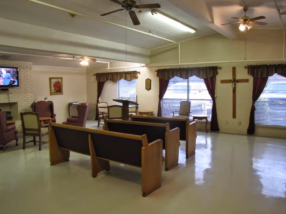Interior room with wooden pews arranged facing a podium, several armchairs, a wall-mounted TV, windows with curtains, ceiling fans, and a large wooden cross on the wall.