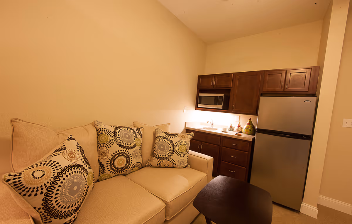 A small living area with a beige couch adorned with three decorative pillows featuring circular patterns. Next to the couch is a compact kitchenette with dark wooden cabinets, a microwave, a small sink, and a stainless steel refrigerator. A small dark wooden table is positioned in front of the couch. The walls are painted a light beige color.