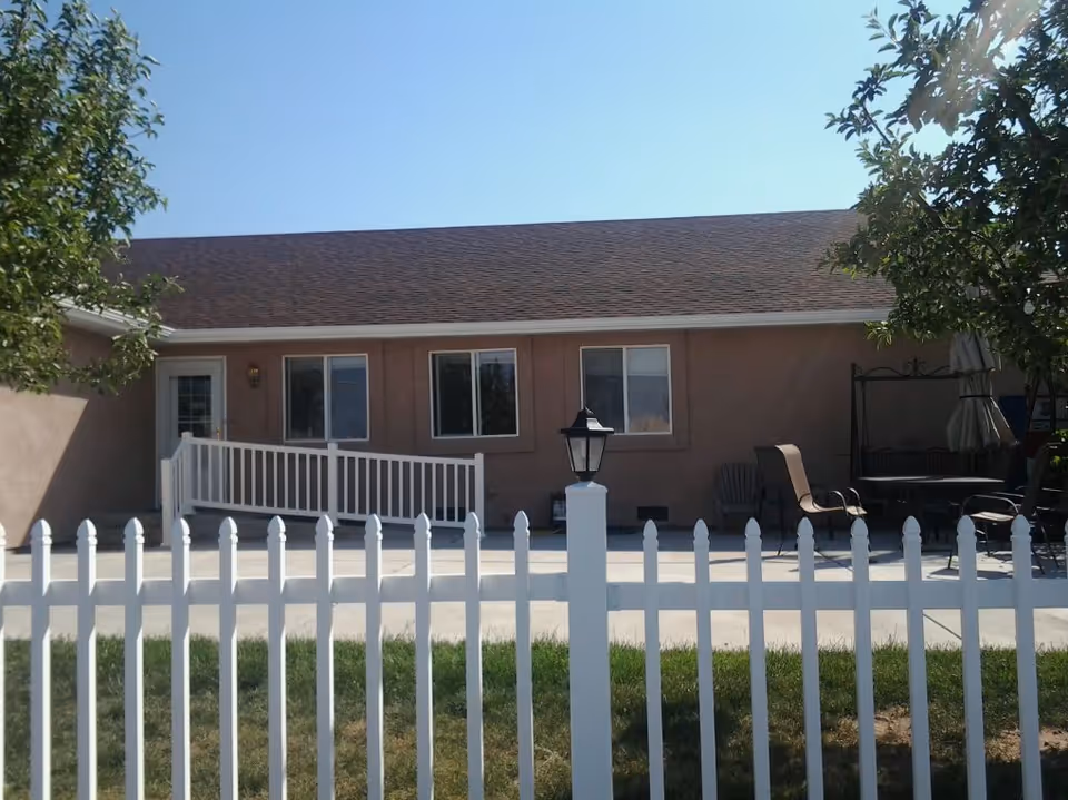 Single-story building exterior with a white picket fence, a concrete patio, ramp to a door, and outdoor seating.