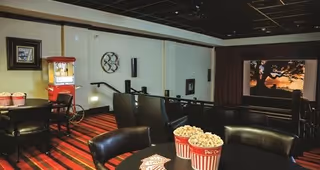 Interior view of a small movie theater room with black leather chairs and tables. On the tables are red and white striped popcorn containers and playing cards. A popcorn machine is visible in the background near the wall, and a movie is playing on a screen showing a tree silhouette against a sunset sky.