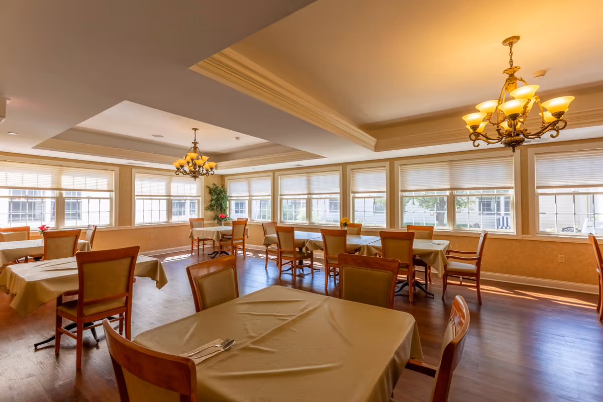 A bright dining room with multiple tables covered in beige tablecloths and wooden chairs. Large windows with blinds allow natural light to fill the room. Two ornate chandeliers hang from the ceiling, and there is a potted plant in the corner.