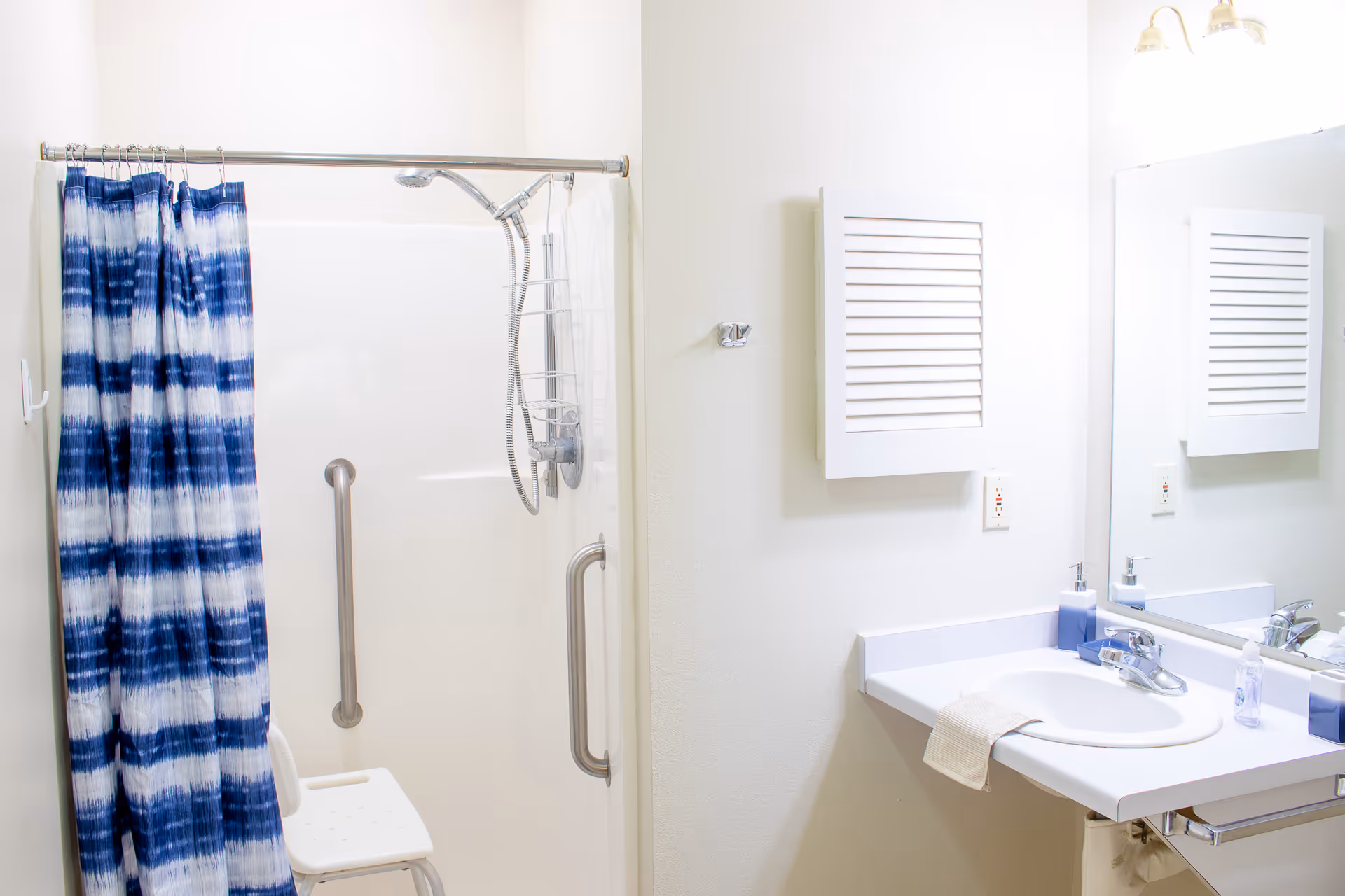 A bright bathroom featuring a white shower stall with a blue and white striped shower curtain, a white shower chair, and a grab bar. To the right is a white sink with a countertop, a mirror above it, a wall-mounted cabinet, and a light fixture with two bulbs.