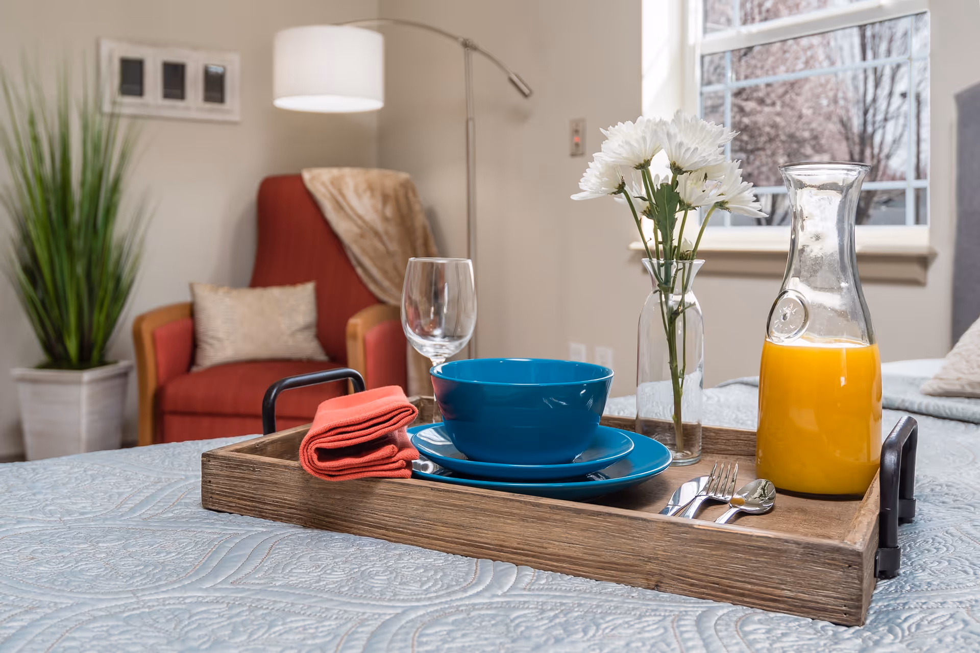 A wooden tray on a bed with a blue bowl and plate, a glass of water, a folded red napkin, silverware, a vase with white flowers, and a glass pitcher of orange juice. In the background, there is a red armchair with a beige pillow and a throw blanket, a tall green plant in a white pot, a floor lamp, and a window showing trees outside.