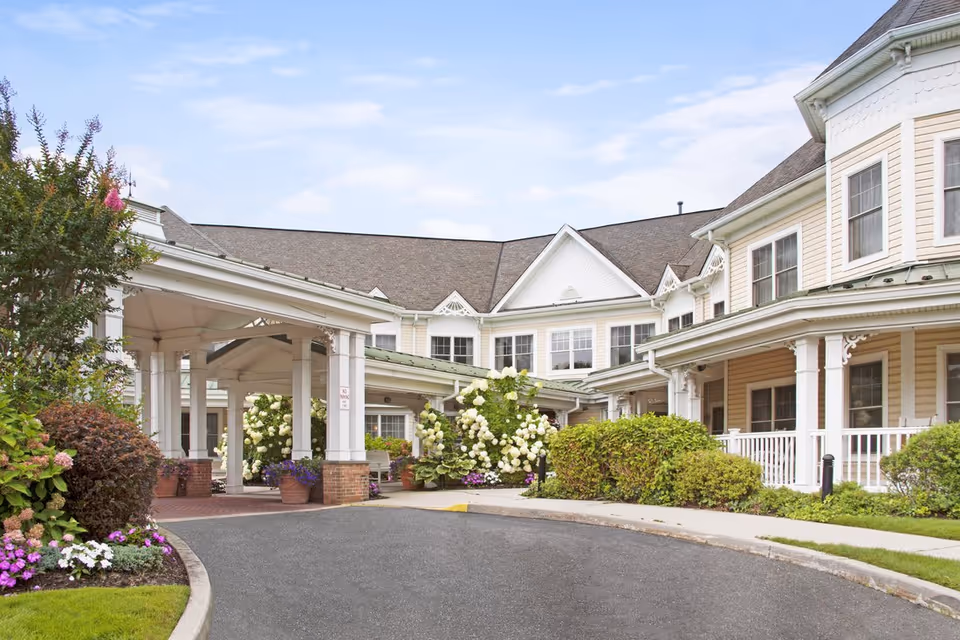 Front entrance of a light-colored senior living building with a covered porte-cochere, potted plants, and flowering shrubs.