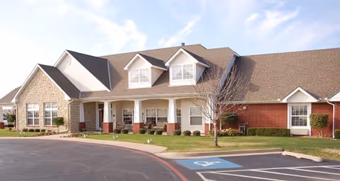 Exterior view of a single-story senior living facility building with a combination of brick and stone facade, a covered entrance with white columns, dormer windows on the roof, and a parking lot with a handicapped parking space in front.