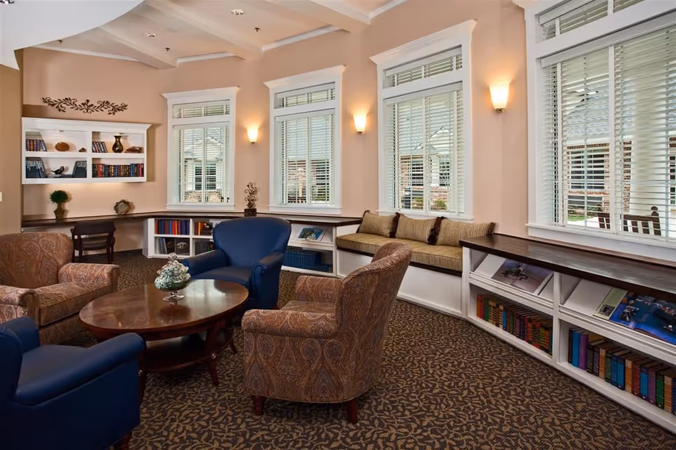 A cozy sitting area in a senior living facility with a round wooden coffee table surrounded by two blue armchairs and two patterned armchairs. The room has large windows with white blinds, built-in bookshelves filled with books and magazines, and a cushioned window bench. The walls are painted a soft beige, and there are wall sconces providing warm lighting.