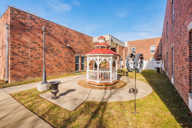 Outdoor courtyard area with a small white gazebo featuring a red roof, surrounded by a circular concrete walkway. The courtyard is enclosed by red brick buildings and has a lamppost, a decorative metal wind spinner, and some planters on the grass.