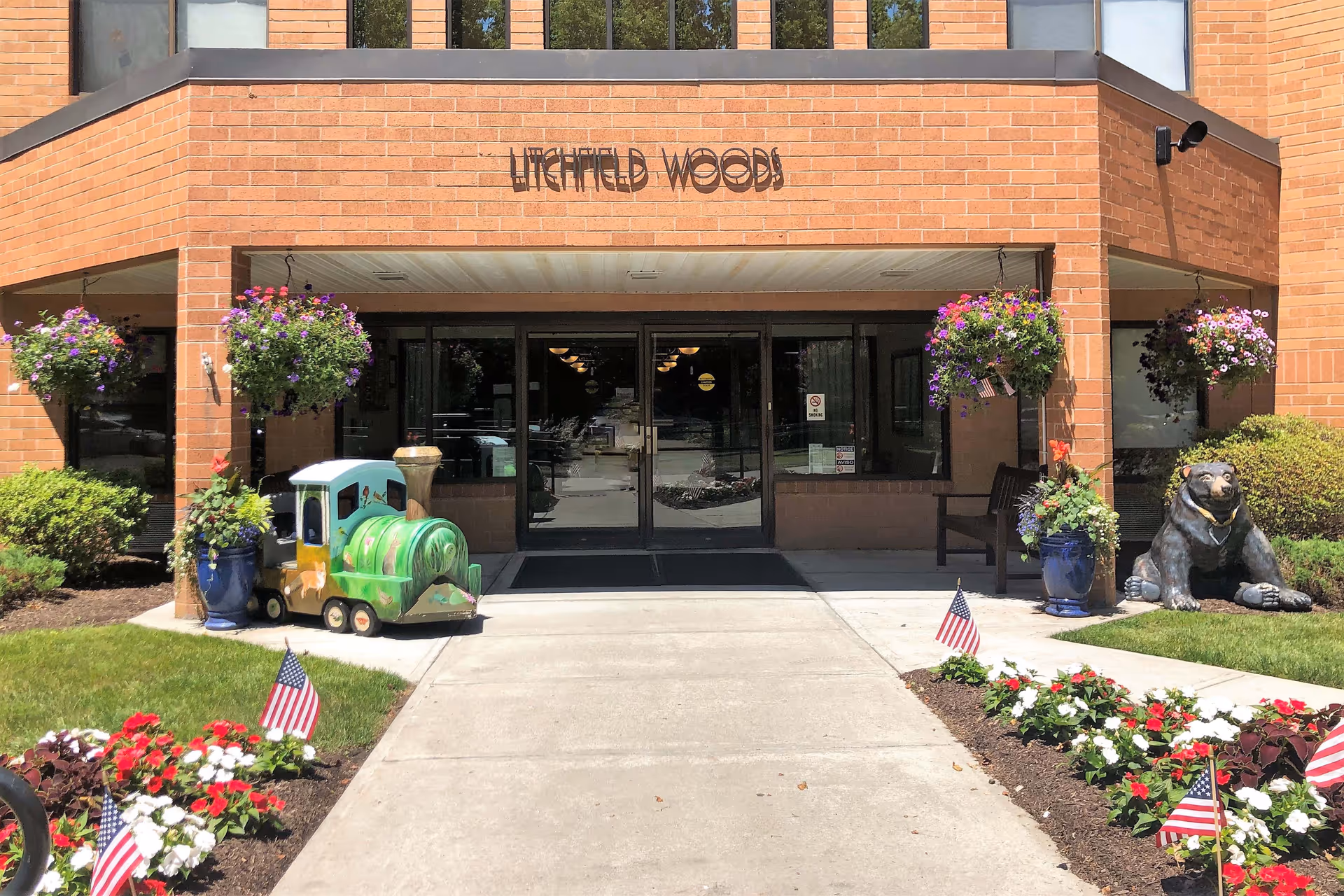 Front entrance of Litchfield Woods with a brick facade, potted flowers, a small children's train ride, American flags, and a bear statue.
