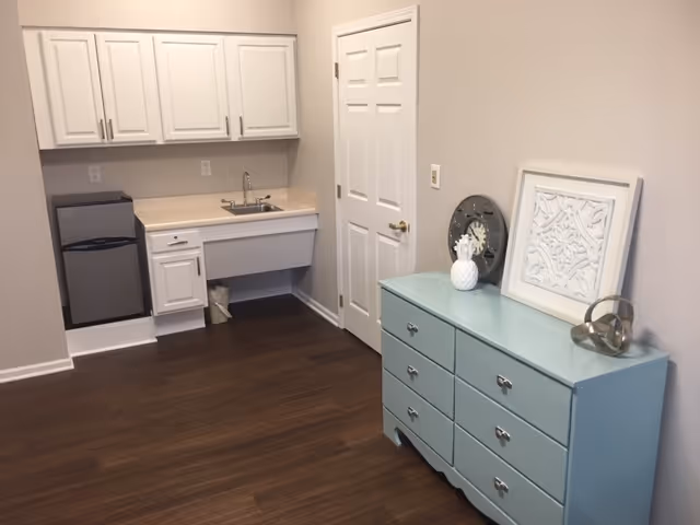 A small kitchenette area with white cabinets, a sink, and a mini refrigerator on the left side. To the right, there is a light blue dresser with decorative items on top, including a framed artwork, a clock, a white pineapple ornament, and a metallic sculpture. The floor is dark wood, and the walls are painted light beige.