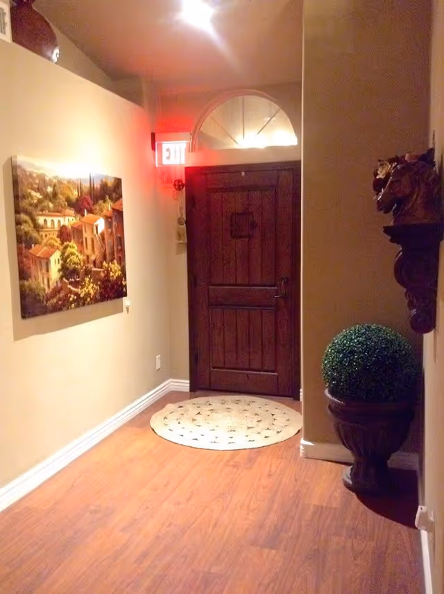 Interior entry hallway with a wooden door beneath a lit EXIT sign, a wall painting, and a decorative potted topiary on hardwood floors.