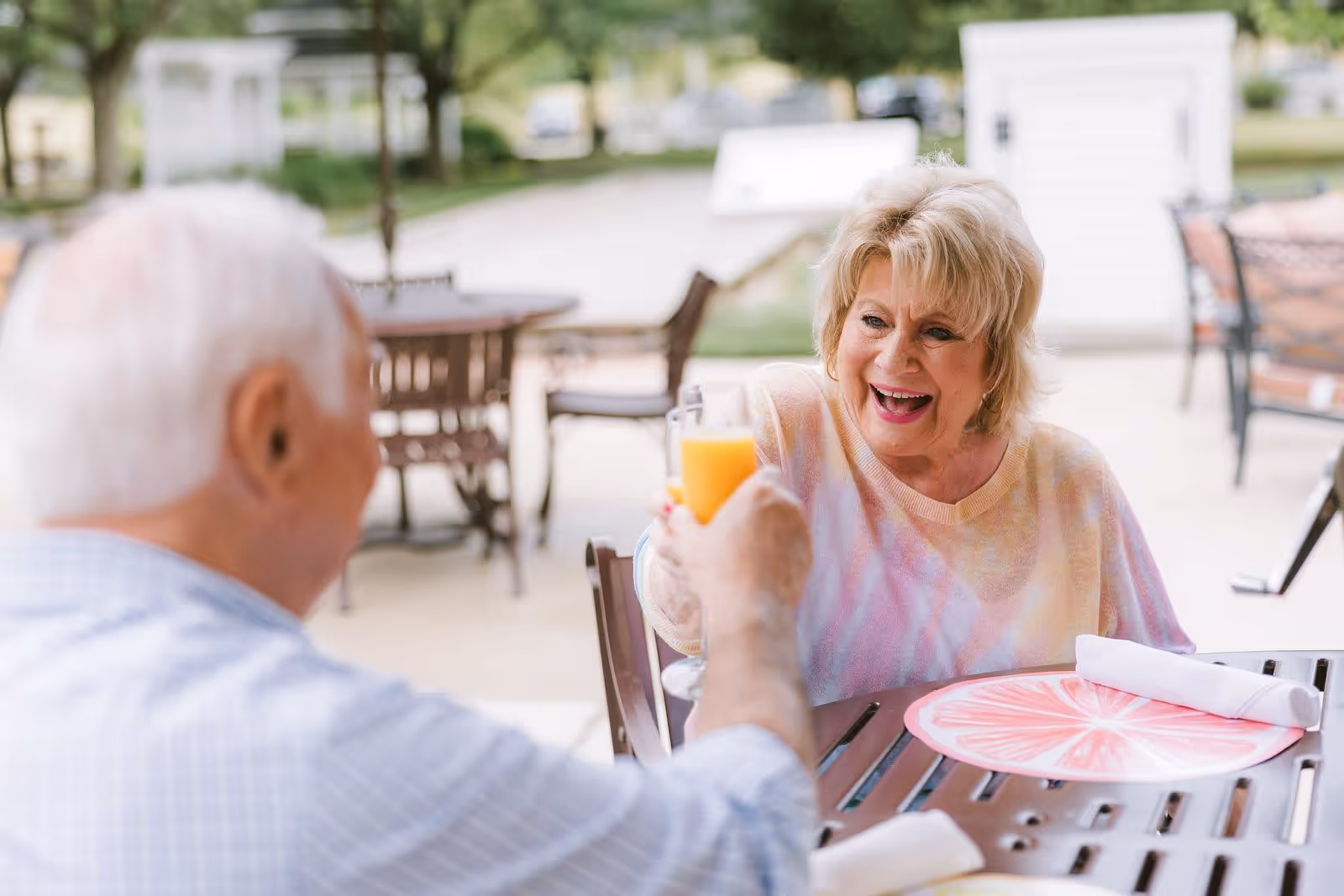 An elderly woman and man sitting at an outdoor table, smiling and clinking glasses of orange juice in a cheerful toast. The setting appears to be a patio area with other tables and chairs in the background.