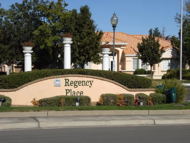 Exterior view of Regency Place Senior Living facility with a beige stucco sign displaying the name Regency Place surrounded by trimmed bushes and trees, with a building and clear blue sky in the background.
