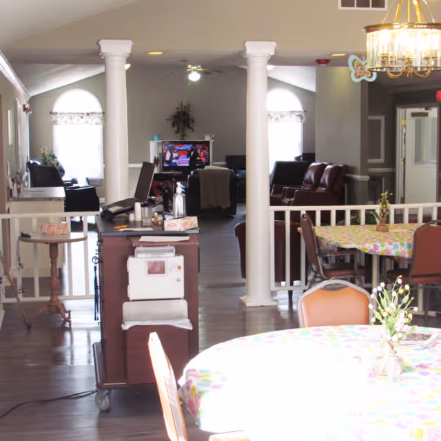 Interior view of a senior living facility common area with round tables covered in colorful tablecloths and chairs around them. In the background, there are white columns, a TV, and several armchairs arranged for seating. The room has large windows with curtains and a chandelier hanging from the ceiling.