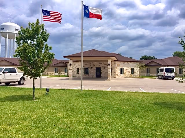 Exterior view of Skilled Care Of Mexia facility showing a single-story stone building with a brown roof, two flagpoles with the American and Texas flags, a green lawn in the foreground, a white pickup truck on the left, a white van on the right, and a water tower in the background.