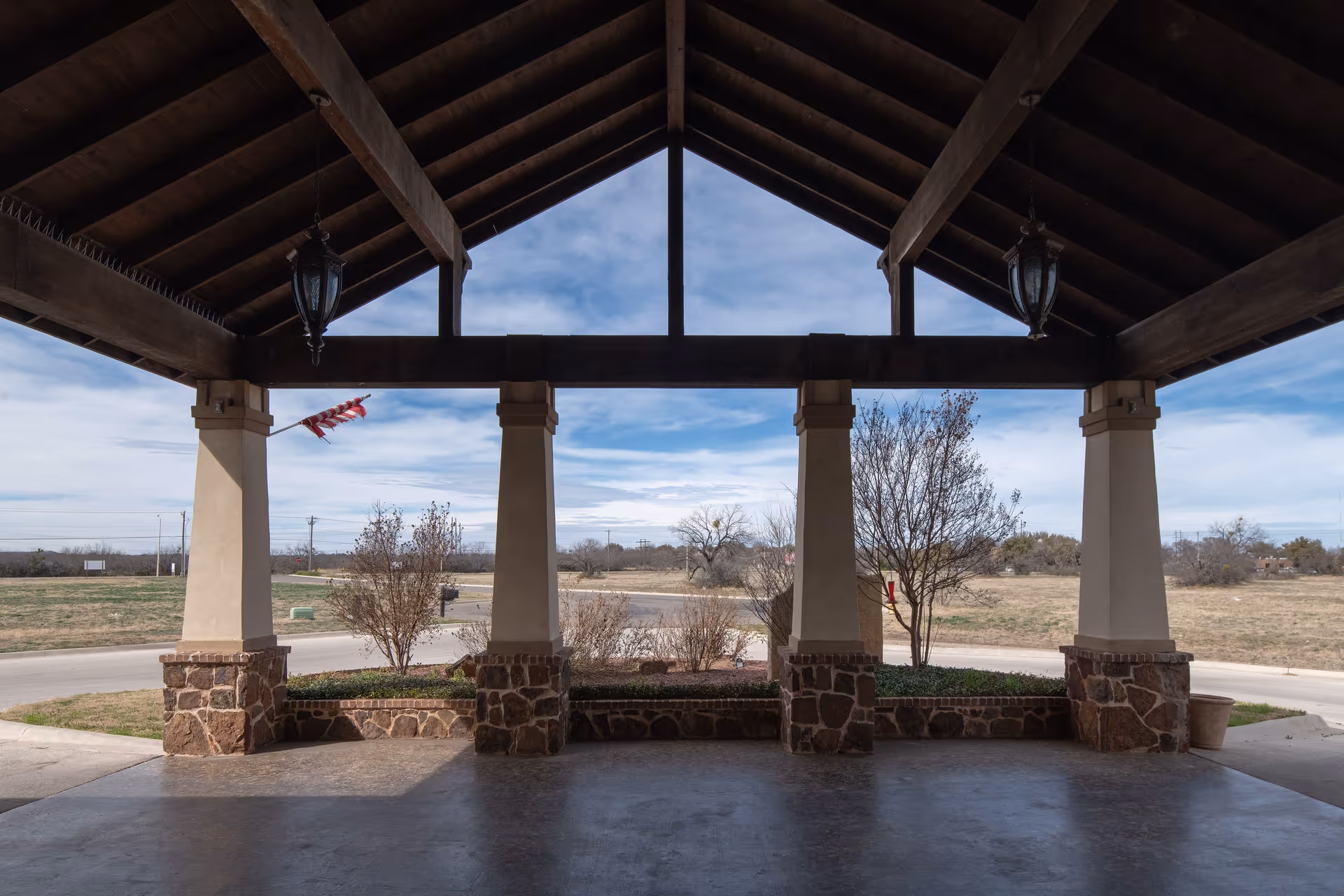 Covered outdoor patio area with a wooden roof supported by stone and stucco pillars, overlooking a landscaped area with small trees and a road beyond under a partly cloudy sky.