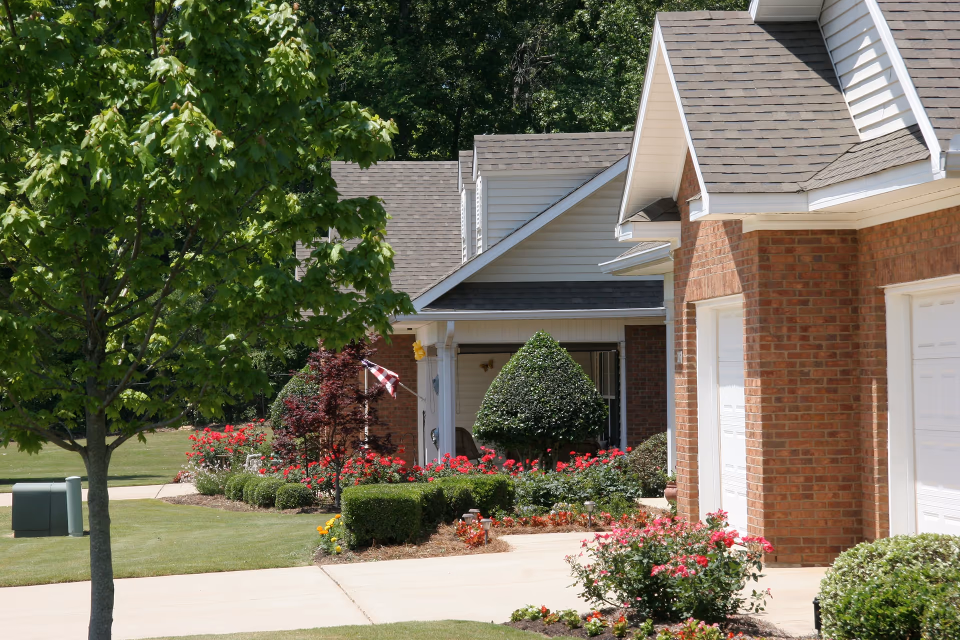 View of a residential area with brick buildings, well-maintained landscaping including trimmed bushes, flowering plants, and a tree. A driveway and sidewalk are visible, along with an American flag near one of the entrances.