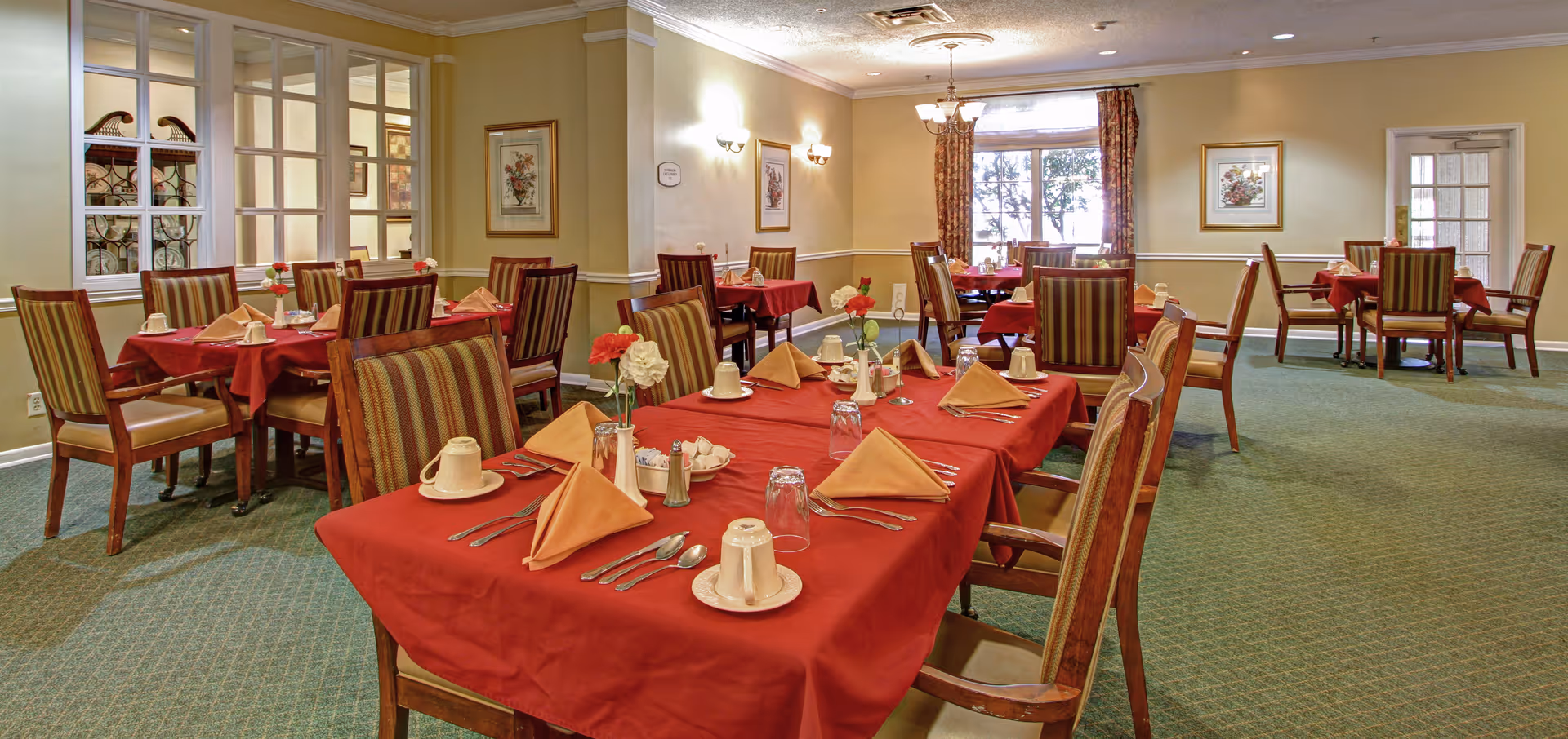 Well-lit dining room with multiple tables covered in red tablecloths, set with folded napkins, cups, and striped chairs.
