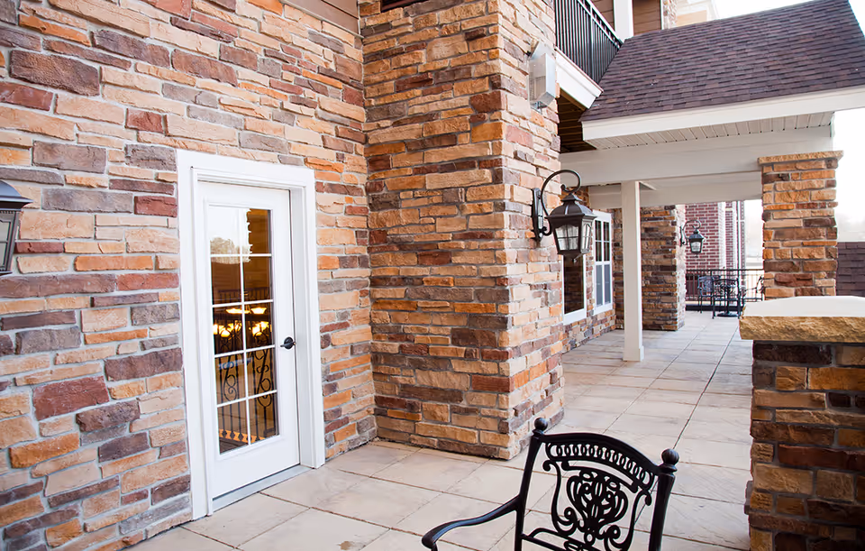 Outdoor covered patio area with stone pillars and walls, a white door with glass panes, black wrought iron chairs, and wall-mounted lantern-style lights.