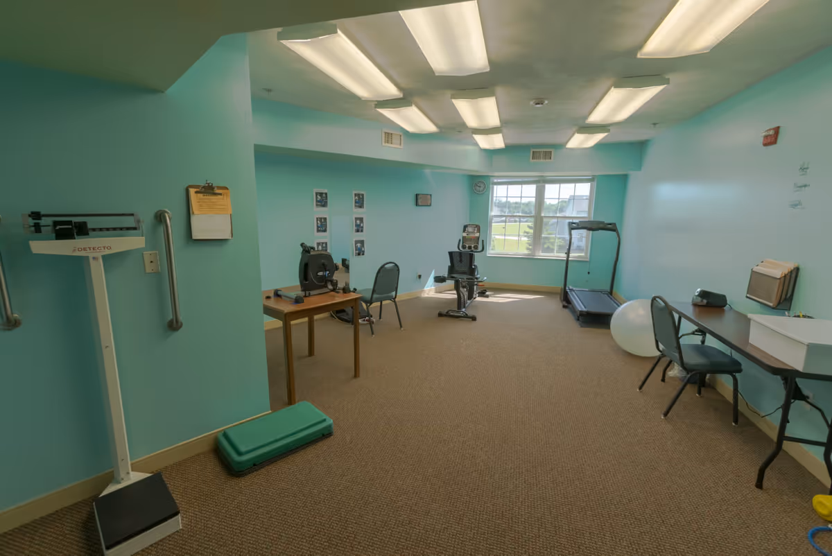 A bright turquoise exercise/therapy room with a scale, step platform, stationary bike, treadmill, exercise ball, tables and chairs.