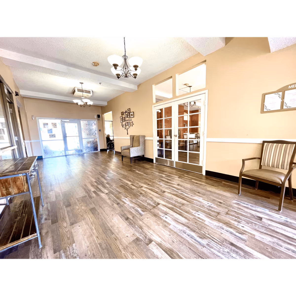 A spacious interior hallway with wood flooring and beige walls. There are two chandeliers hanging from the ceiling, a wooden console table on the left, and several chairs along the walls. A set of glass double doors leads to another room, and a wall decoration with the word 'family' is visible. A person in a wheelchair is near the far end of the hallway by the entrance doors.