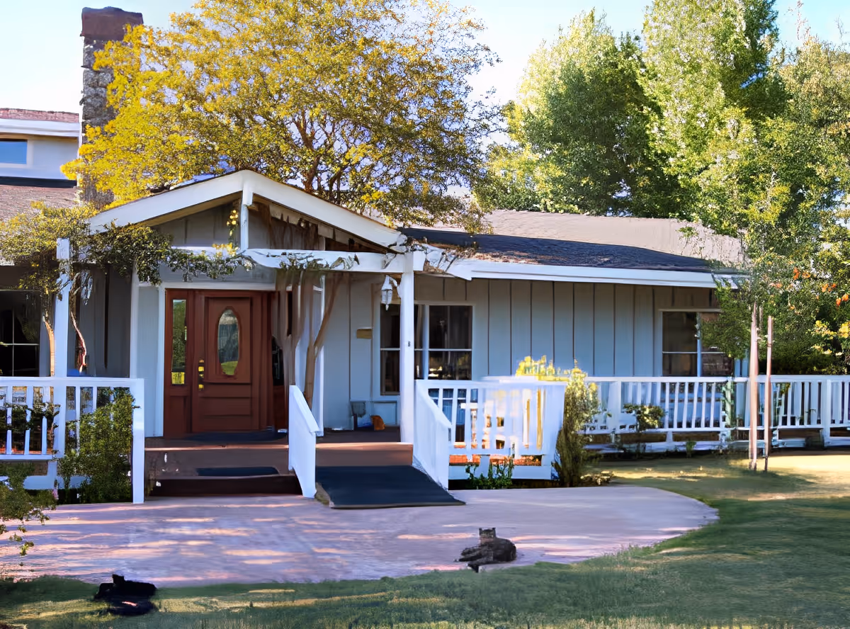 Exterior view of a single-story house with a white wooden porch and a ramp leading to a red front door. The house is surrounded by green trees and a well-maintained lawn with two cats lying on the grass near the porch.