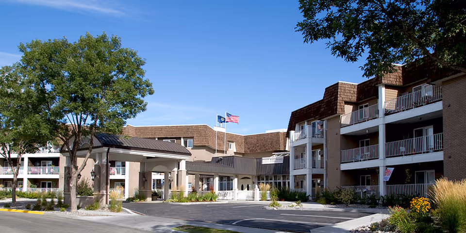 Front exterior of a senior living building with a covered entrance/porte-cochère, balconies, landscaping, and flagpoles.