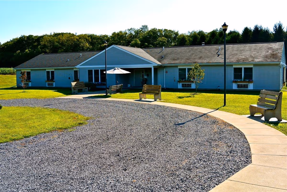 Single-story building with light blue siding and a gray roof, surrounded by a green lawn and trees in the background. There is a curved gravel driveway and a concrete walkway with wooden benches and street lamps along the path. An umbrella is visible near the entrance of the building.