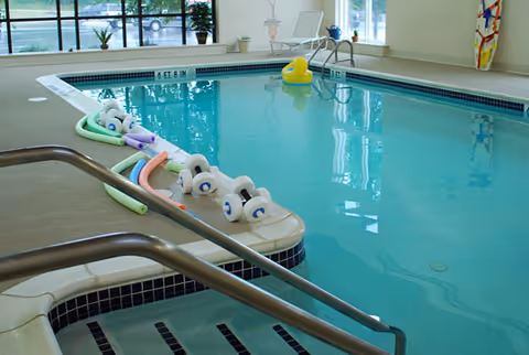 Indoor swimming pool with clear blue water, pool noodles and foam dumbbells floating on the surface, metal handrails leading into the pool, large windows letting in natural light, and a yellow rubber duck float near the far end.