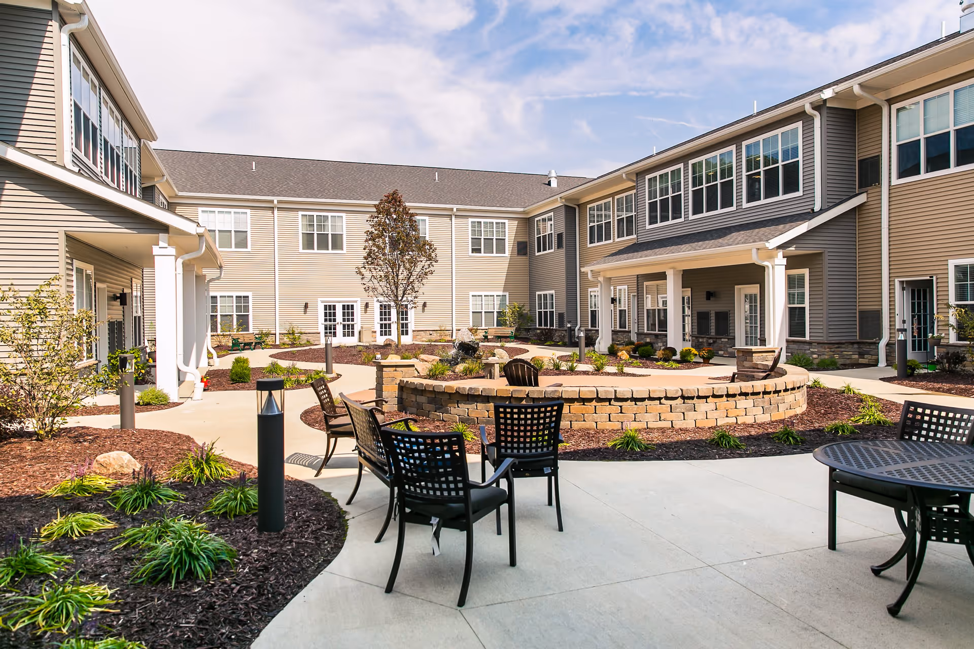 Outdoor courtyard area of a senior living facility with a circular brick fire pit surrounded by black metal chairs and tables. The courtyard is landscaped with small plants and trees, and the building has beige siding with many windows under a partly cloudy sky.