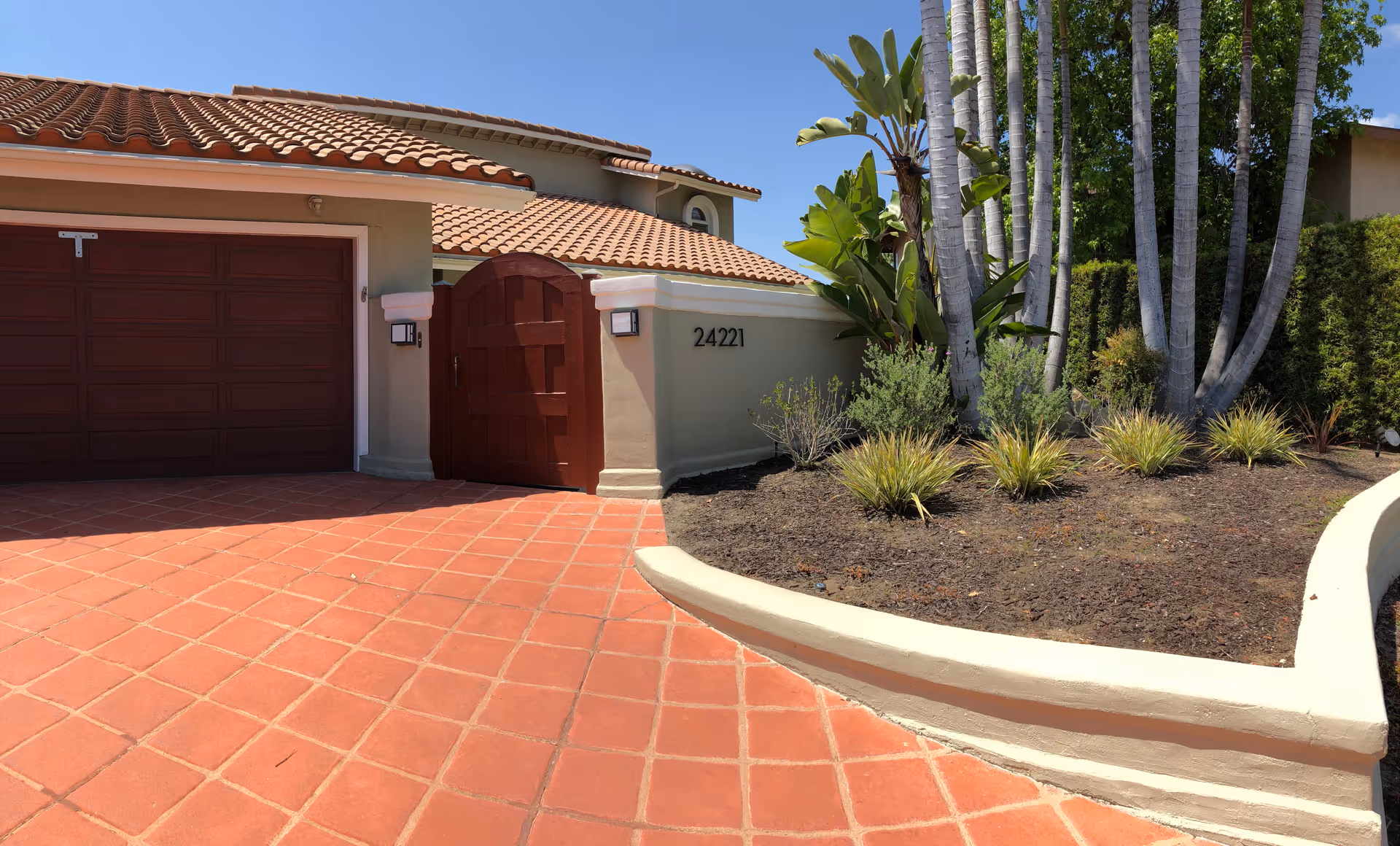 Exterior view of a residential building with a red tiled driveway, a closed wooden garage door, a wooden gate, and a garden area with various plants and tall palm trees. The building has a beige stucco finish and a red tile roof under a clear blue sky.