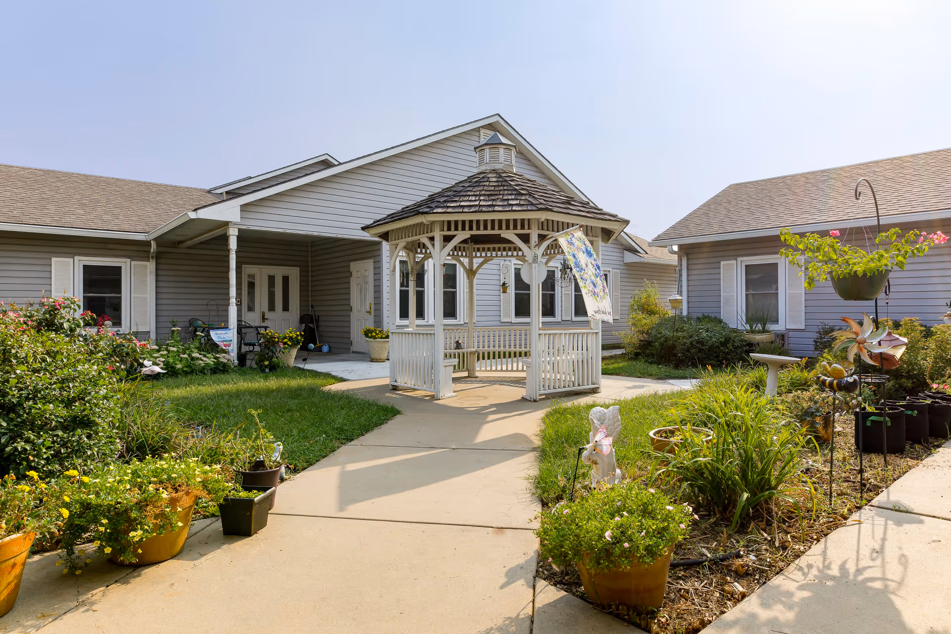 Outdoor courtyard area of Homestead Assisted Living of Derby featuring a white wooden gazebo in the center, surrounded by green plants, flowers in pots, and garden decorations. The courtyard is bordered by single-story buildings with gray siding and white trim under a clear sky.