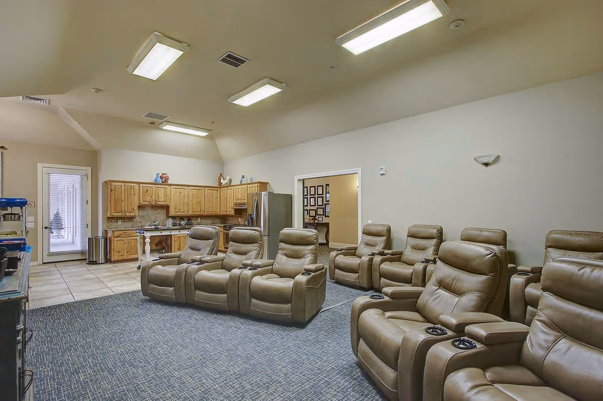 A spacious room with multiple beige leather recliner chairs arranged in two rows facing a TV or entertainment area. The room has a carpeted floor with a tiled section near a kitchen area featuring wooden cabinets, a stainless steel refrigerator, and a door leading outside. The ceiling has several rectangular fluorescent lights.