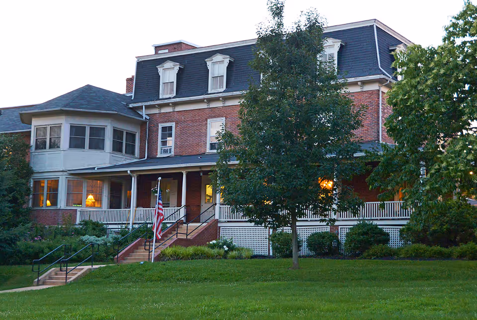 A large, multi-story brick building with a dark mansard roof and white trim, surrounded by green trees and a well-maintained lawn. The building has a porch with white railings and steps leading up to the entrance, where an American flag is displayed. Warm lights are visible through some windows, indicating interior lighting.